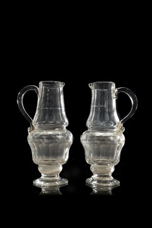 Coppia di ampolle in vetro incolore decorate a sfaccettature, anse sagomate. In