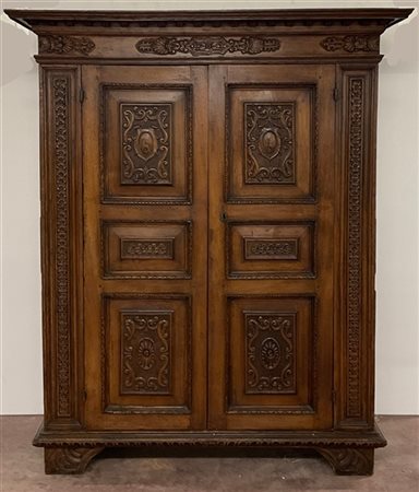 Armadio in noce a due ante pannellate con cappello aggettante, due decori a intaglio a stemmi entro volute e due a motivi fioriti. Fianchi pannellati. Lombardia, inizio secolo XVIII (cm 163x214x64) (difetti, restauri e intagli di epoca posteriore)