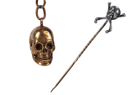 Lotto composto da un pendente porta pillole in foggia di teschio con mandibola apribile in oro giallo 18K (g 10; cm 2) ed uno spillone fermacravatta in oro giallo 18K sormontato da teschio con ossa incrociate in argento (g 9; cm 8) 