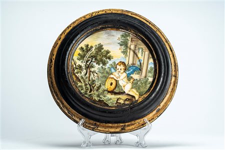 Piattello in maiolica policroma istoriata raffigurante putto alato con tamburo in un paesaggio agreste con rovine classiche. Entro cornice lignea. Att […]