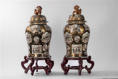 Coppia di potiche in porcellana decorate con scene di vita quotidiana entro pannelli su fondo nero con oro, coperchio con shishi, Kutani, Giappone. Basi in legno, 20th century